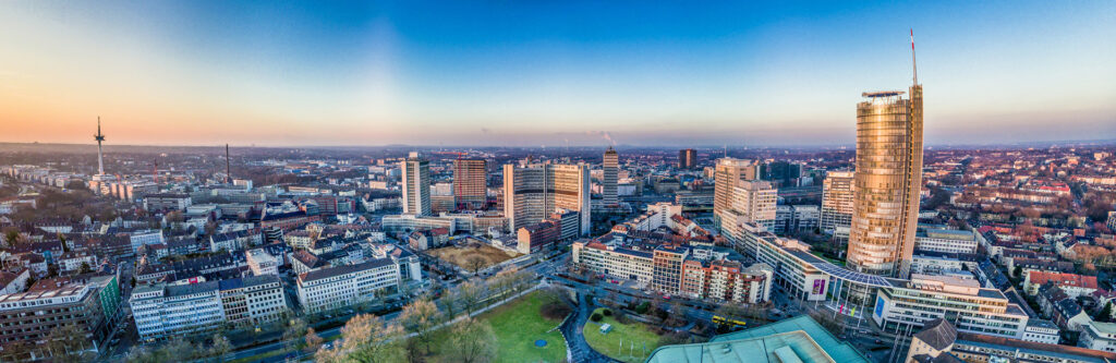 Foto für die Standortseite "Schuldnerberatung Schulz in Essen". Das Bild zeigt die Skyline von Essen bei Sonnenuntergang. Im Vordergrund erkennt man den modernen Bürokomplex mit dem markanten RWE-Turm, der das Stadtbild prägt. Die Aufnahme bietet einen weiten Blick über die dicht bebaute Innenstadt und den umliegenden Ruhrpott – ein Mix aus moderner Architektur, Industrie und urbanem Leben.