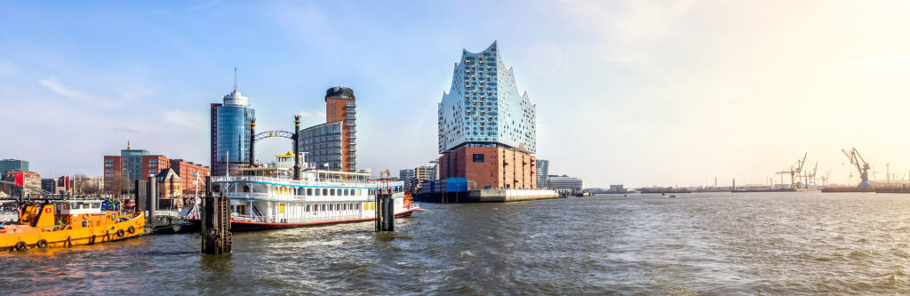 Foto für die Standortseite "Schuldnerberatung Schulz in Hamburg". Das Bild zeigt den Hamburger Hafen mit der markanten Elbphilharmonie im Mittelpunkt. Die moderne Architektur des Konzerthauses ragt über dem historischen Backsteinsockel empor. Im Vordergrund liegt ein nostalgisches Schaufelradschiff vertäut, während rechts die Hafenkräne die industrielle Seite der Stadt unterstreichen. Die Szene verbindet Tradition, Moderne und wirtschaftliche Bedeutung.