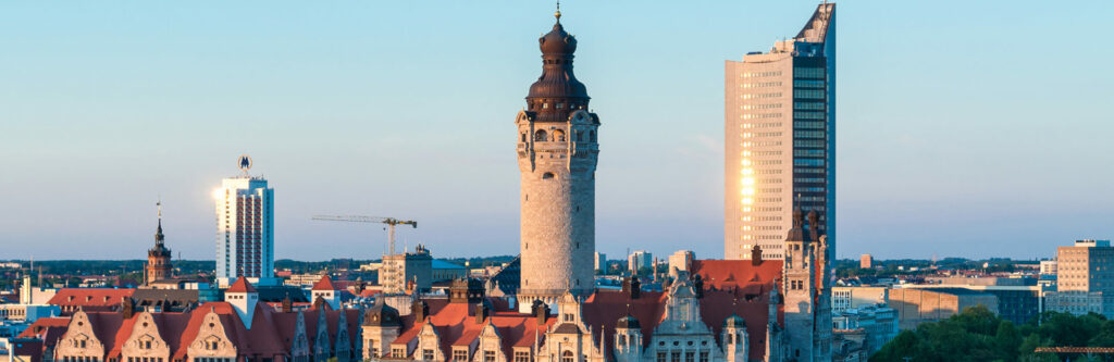 Foto für die Standortseite "Schuldnerberatung Schulz in Leipzig". Das Bild zeigt die Skyline von Leipzig mit einer Mischung aus historischer und moderner Architektur. Im Vordergrund ragt das Neue Rathaus mit seinem markanten Turm empor. Dahinter ist das City-Hochhaus zu sehen, eines der höchsten Gebäude der Stadt. Die Szene ist in warmes Licht getaucht, vermutlich während des Sonnenuntergangs.