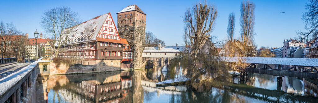 Foto für die Standortseite "Schuldnerberatung Schulz in Nürnberg". Das Bild zeigt eine malerische Winteransicht der Altstadt von Nürnberg. Zu sehen ist das berühmte Heilig-Geist-Spital mit seinem markanten Fachwerkbau direkt am Pegnitzufer, daneben die alte Stadtmauer mit dem Wehrturm. Der Schnee verleiht der Szene eine ruhige, fast märchenhafte Atmosphäre.