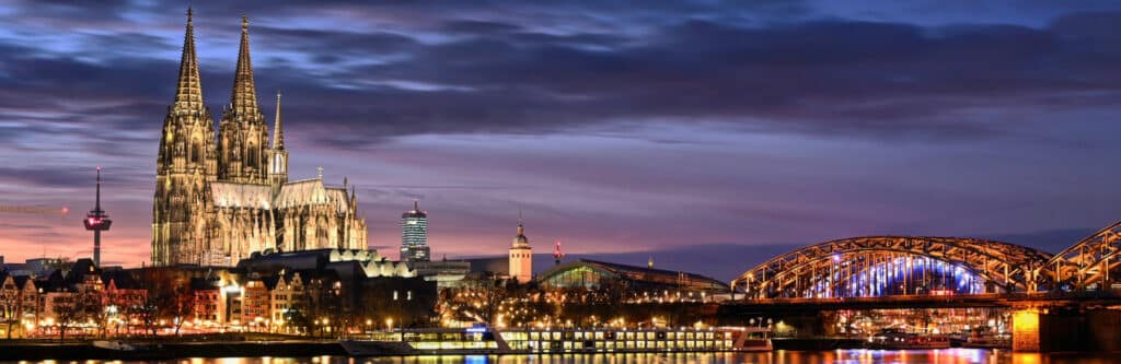 Foto für die Standortseite "Schuldnerberatung Schulz in Köln". Das Bild zeigt den berühmten Kölner Dom bei Abenddämmerung, daneben die Hohenzollernbrücke über den Rhein und das Lichterspiel der Stadt Köln.