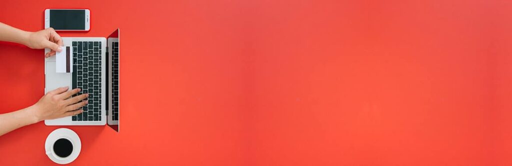 Foto für den Blogartikel "Kann ich im Online-Banking eine Pfändung sehen?". Blick von oben auf ein Notebook, Hand mit Karte, daneben Smartphone und Kaffee auf rotem Hintergrund – Sinnbild für Online-Banking und das Prüfen, ob eine Kontopfändung im Konto sichtbar ist.