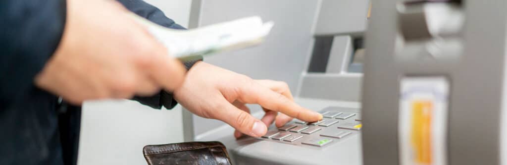 Foto für den Blogartikel "P-Konto: Wann kann ich über das Geld verfügen?". Auf dem Foto: Person gibt an einem Geldautomaten eine Eingabe über die Tastatur ein und hält Banknoten in der Hand – Symbolbild für die Verfügbarkeit von Guthaben auf dem P-Konto nach Zahlungseingang oder Freigabe.